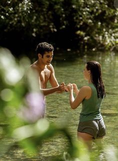 a man and woman standing in the water holding each other's hands with trees in the background