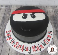 a black and white birthday cake with a ninja face on it's top is sitting on a wooden table