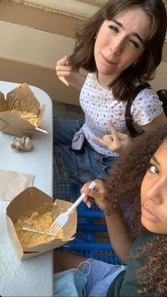 two women sitting at a table with food in boxes on the table and one holding a fork