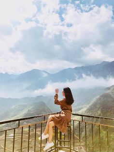 a woman sitting on top of a metal railing next to mountains and clouds in the sky