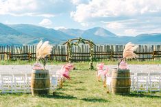 an outdoor ceremony set up with white chairs and wooden barrels filled with pamodia