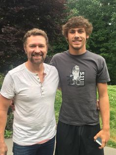 two men standing next to each other in front of green grass and trees on a sunny day