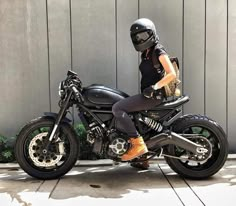 a woman sitting on top of a black motorcycle next to a gray wall and wearing a helmet