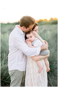 a man and woman holding a baby in their arms while the sun sets behind them