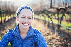 a woman in a blue jacket smiles at the camera while standing next to rows of vines