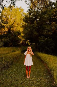 a girl in a white dress is standing on the side of a dirt road with trees and grass behind her