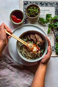 two hands holding chopsticks over a bowl of noodles with meat and vegetables on the side