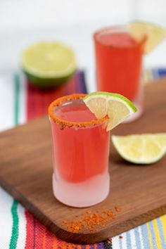 two glasses filled with red liquid and limes on a wooden cutting board next to a striped towel