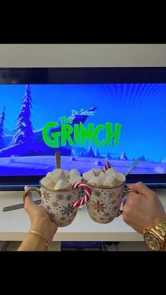 two people are holding mugs with marshmallows in front of a tv