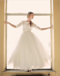 a woman in a white dress standing by a window with her hands out to the side