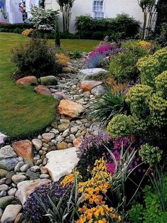 a garden with rocks and flowers in front of a white house on the side of a hill