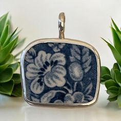 a blue and white flowered square pendant on a silver chain next to succulents