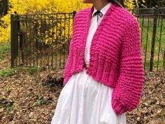 a woman wearing a pink knitted cardigan and white dress standing in front of a fence