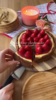 a person cutting into a strawberry cheesecake on top of a wooden table next to a candle