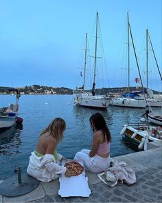 two women are sitting on the dock eating pizzas and watching boats in the water