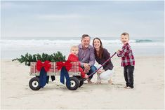 a man and woman with two children on the beach next to a wagon that has a christmas tree in it