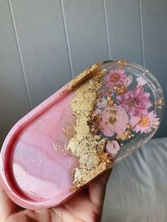 a hand holding a pink and gold tray with flowers on it in front of a white wall