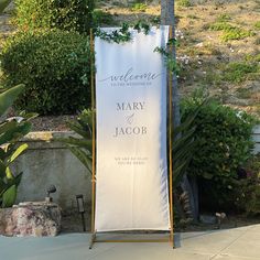 a welcome sign with greenery on it in front of some bushes and trees at a wedding