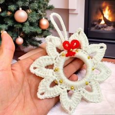 a hand holding a crocheted ornament next to a christmas tree with a fireplace in the background