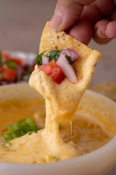 a hand holding a tortilla chip over a bowl of soup with tomatoes and onions