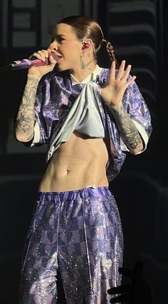 a woman with tattoos on her chest singing into a microphone while wearing purple pants and a silver crop top