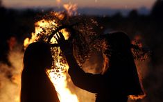 two women standing in front of a fire with their hands on each other's shoulders