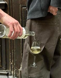 a man pouring white wine into a glass in front of an open door with another person standing next to him