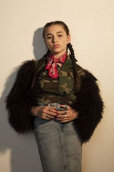 a woman wearing a camouflage shirt and black fur coat standing in front of a white wall