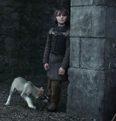 a young boy standing next to a dog on top of a dirt ground near a stone wall