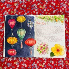 an open notebook with watercolors and paper lanterns on the cover, sitting on a red flowered tablecloth