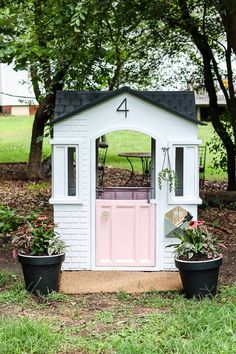 a small white shed with pink door and windows in the grass next to some potted plants