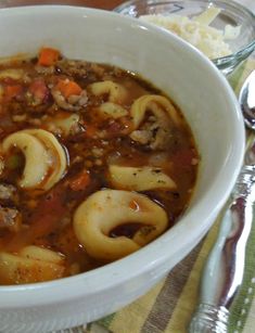 a white bowl filled with pasta and meat soup on top of a table next to silverware