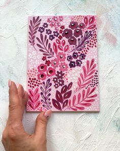 a hand is holding up a pink and purple painting on a white surface with flowers