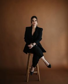 a woman sitting on top of a wooden stool in front of a brown wall with her legs crossed