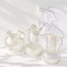 three white vases sitting next to each other in front of a drawing on the wall