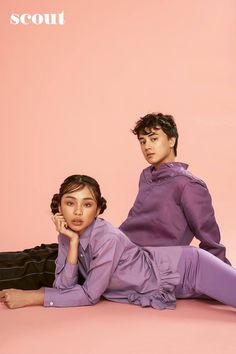 two young people sitting on the ground posing for a photo in front of a pink background