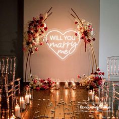 a wooden table topped with lots of candles and flowers next to a neon sign that says will you marry me?