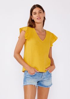 [Color: Mustard] An image of a brunette model wearing a mustard yellow top crafted from cotton gauze. With short flutter sleeves and v neckline. A comfortable top for the summer to fall season that can be paired with denim shorts and pants. Casual Cotton Top With Butterfly Sleeves, Casual Cotton Tops With Butterfly Sleeves, Flutter Sleeve Tops For Summer Brunch, Ruffled Cotton Short Sleeve Top, Summer Tops With Flutter Sleeve For Brunch, Ruffle Sleeve Cotton Top With Ruffles, Cotton Short Sleeve Top With Ruffle Sleeves, Casual Butterfly Sleeve Blouse For Summer, Spring Short Sleeve Cotton Gauze Tops