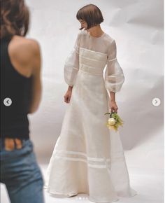 a woman standing in front of a white backdrop wearing a dress and holding a flower