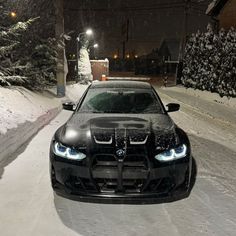 a black car parked on the side of a snow covered road at night with its lights on