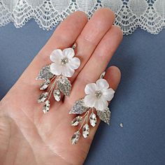 a pair of white flower earrings with leaves and crystals in the shape of flowers, on a woman's hand