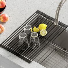 three glasses and two lemons on a kitchen sink rack next to some apples in a bowl