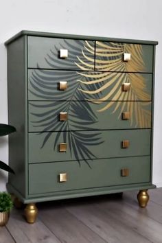 a green dresser with gold leaves painted on the front and sides, next to a potted plant