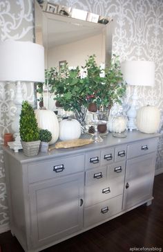 a silver dresser topped with lots of white vases and plants next to a mirror