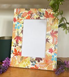 an empty photo frame sitting on top of a wooden table next to flowers and a cup