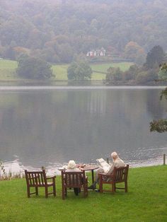 two people sitting at a table on the grass near a body of water with trees in the background