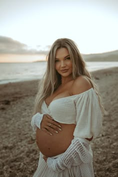 a pregnant woman posing on the beach with her belly wrapped around her waist and wearing an off - shoulder white top