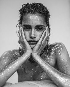 a woman with her hands on her face and body covered in silver glitter, looking at the camera