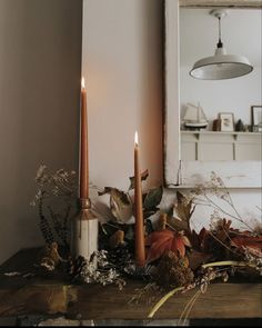 two candles sitting on top of a wooden table next to leaves and flowers in front of a mirror