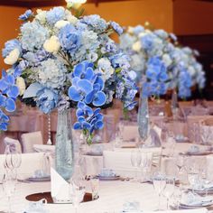 blue and white flowers are in tall vases on the dining room table set for an event